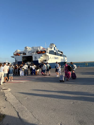 A group of people are disembarking from a large white ferry with the word "SEAJETS" visible on its side. The ferry is docked at a port, and there is a clear blue sky overhead. The people are dressed in casual summer clothing and are carrying luggage, including suitcases and backpacks. Some are walking towards the camera, while others are walking away. The ground is paved and has red and white striped markings, possibly indicating a pedestrian crossing. The ferry has several decks and life rafts are visible along its side. In the background, the sea is visible, and there is another smaller ferry or boat in the distance. The overall scene suggests a busy port at sunset or sunrise, with travelers arriving or departing. The location is the port of Heraklion, Greece, evident from the context provided with the prompt.