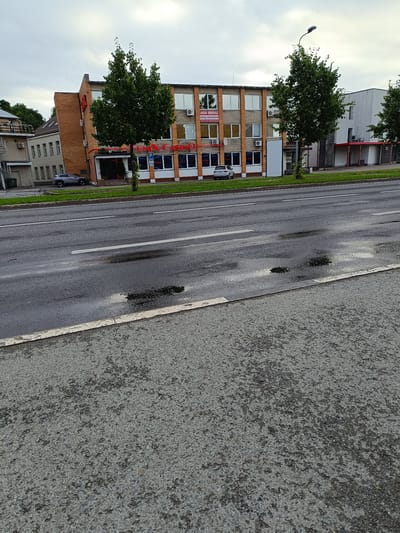 The image shows a street scene in Jõhvi, Estonia. On the right side of the street is a building with a brick facade, featuring large windows with the text "FENIX CASINO" in red lettering. Above the windows, there are several air conditioning units. Adjacent to the casino building is a white building with red awnings. Trees line the sidewalk in front of the buildings. The street itself appears wet, with several puddles and reflections on the asphalt. A gray car is parked on the street in front of the casino. The sky is overcast.