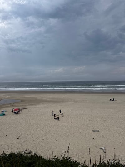 An overcast sky looms above a wide, sandy beach in Arch Cape, Oregon, United States. The ocean stretches to the horizon, with several waves breaking on the shore. In the mid-ground, a group of people are gathered under a red umbrella near a tent, possibly having a picnic or enjoying the beach. Further to the right, another smaller group is visible, and a lone person can be seen walking near the water. The sand is dotted with driftwood and a few small puddles. In the foreground, tall grasses and foliage line the edge of the beach, obscuring a fire pit.