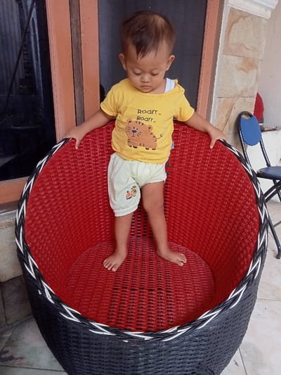 A young child stands in a large, round, woven chair. The chair is black and red with white trim. The child wears a yellow t-shirt with a tiger design and light-colored shorts. The child is looking down with their hands resting on the chair's rim. In the background, there's a doorframe, a wall, and a blue chair. The scene appears to be outdoors on a tiled surface.