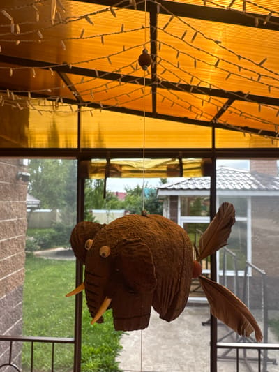 An elephant-shaped decoration, likely made from coconut husk, hangs suspended by a string from an overhead structure with orange translucent panels and white string lights. The elephant decoration has large googly eyes, tusk-like protrusions, and feathers attached to its rear. It is positioned in front of a glass-paned doorway or window, with a blurred outdoor scene visible behind it. The outdoor scene shows green grass, trees, houses with tiled roofs, and a stone-clad wall on the left. The setting appears to be a porch or covered patio. The photograph was taken in Chesnokovka, Russia.
