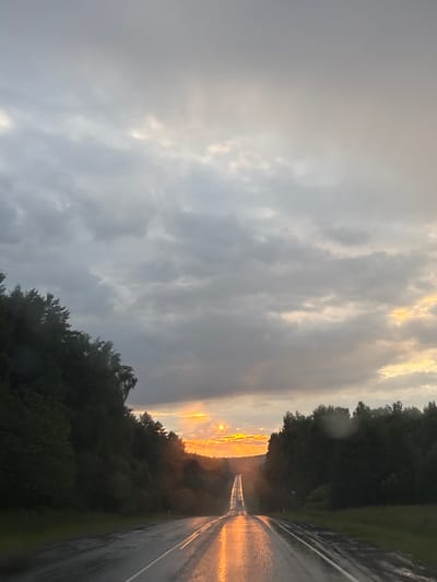 The image depicts a wet, paved road in Malyi Dubovik, Russia, stretching into the distance under a dramatic, overcast sky. The sun is setting, casting a bright orange glow on the clouds and reflecting intensely on the wet asphalt, creating a shimmering effect. Lush green trees line both sides of the road, their foliage dark against the moody sky. A single vehicle is visible on the road in the distance. The overall atmosphere is one of a rainy, yet beautiful, evening.