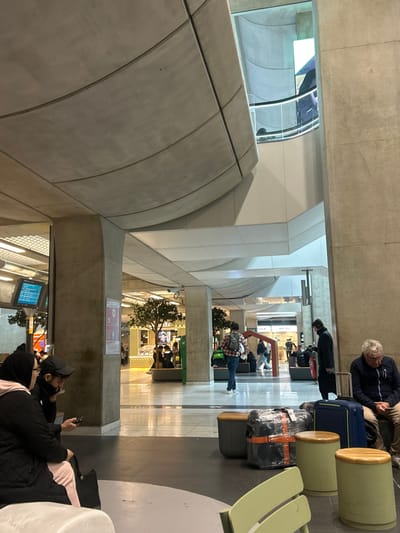 This image depicts an interior concourse of a modern multi-level transit terminal, identified as Charles de Gaulle Airport in Tremblay-en-France, France. The environment features a polished light-colored tiled floor with integrated darker circular patterns, large concrete support columns, and a ceiling composed of curved, segmented concrete panels. An upper level with a glass railing is visible in the background, suggesting multiple floors.
In the immediate foreground left, two individuals are seated on a light-colored bench. One individual, on the left, wears a black hijab and a black quilted jacket over light pink attire. The second individual, to the right, wears a black baseball cap and a dark jacket, and is observed looking at a mobile phone.
In the mid-ground right, an older male with grey hair, wearing a dark blue zip-up jacket, is seated on a light green bench, looking downwards. Next to him are a dark blue hard-shell suitcase and a large black, plastic-wrapped bundle secured with orange tape. To his left, a male wearing a dark coat and headphones stands upright, positioned near additional dark luggage.
Further into the mid-ground, a male wearing a plaid shirt and a backpack walks away from the viewer, carrying a bright green duffel bag. Beyond these individuals, the background reveals various retail or food service establishments, some featuring decorative artificial trees and warm interior lighting. Multiple other individuals are visible throughout the concourse, engaged in walking or sitting on grey benches. Information display screens are mounted on a concrete pillar in the mid-ground left. The overall lighting is artificial and evenly distributed throughout the large open space.