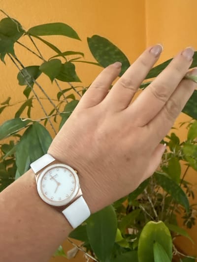 A close-up shot captures a person's left wrist adorned with a white-strapped watch featuring a white face and rose-gold accents. The watch has small, diamond-like markers at each hour position. The person's hand is slightly raised, and their fingers are visible, with light-colored nail polish. In the background, out-of-focus green leaves from a plant are visible against an orange wall. The scene appears to be indoors. There is no information available about the location in Hraniv, Ukraine.