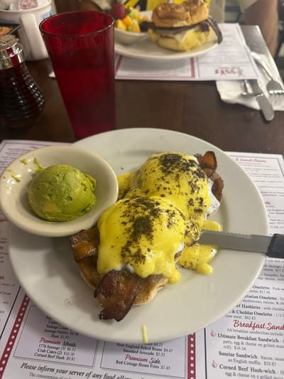 The image shows a close-up of a breakfast meal on a plate, set on a table in what appears to be a restaurant. The meal consists of two eggs benedict with bacon and a side of mashed avocado. The eggs are covered in hollandaise sauce and sprinkled with herbs. A knife is positioned to the right of the plate. In the background, a red glass of beverage, a syrup dispenser, and a partially visible sandwich are present. A menu with various breakfast and lunch options is visible beneath the plate. The scene suggests a casual dining setting, likely a diner or cafe. The resolution of the image makes it difficult to determine specific details about the people present or the exact location within the city of Dennis.
