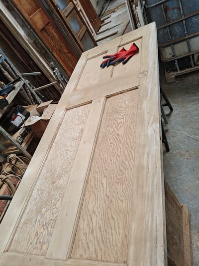 A stripped, pale wooden four-panel door rests horizontally on dark sawhorses in a workshop environment in Swindon, United Kingdom. A pair of red and black work gloves lies on the upper rail of the door. The door's surface displays prominent light wood grain, characteristic of having been sanded or stripped of finish. The background to the left features cluttered metal shelving holding stacked wooden panels, some dark, others lighter, alongside power tools with coiled cables and an open cardboard box containing white, irregular items. To the upper right, a large, dark grey industrial bulk container (IBC) with a metal cage framework is visible. The concrete floor in the foreground and surrounding areas appears dusty and stained. No individuals are present.