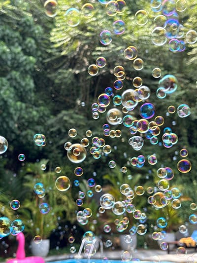 The image depicts an outdoor scene in Unawatuna, Sri Lanka. Hundreds of iridescent soap bubbles of varying sizes, displaying prismatic colors, are suspended across the frame. The background, rendered in soft focus, consists of dense green foliage, including large trees and tropical shrubs. In the lower portion of the image, a bright blue swimming pool is partially visible, containing a pink inflatable flamingo positioned in the lower-left. A water feature, possibly a small fountain, is present within or beside the pool on the right. Additional potted plants with green leaves are situated around the pool's edge. The scene is illuminated by natural daylight. No individuals are present.