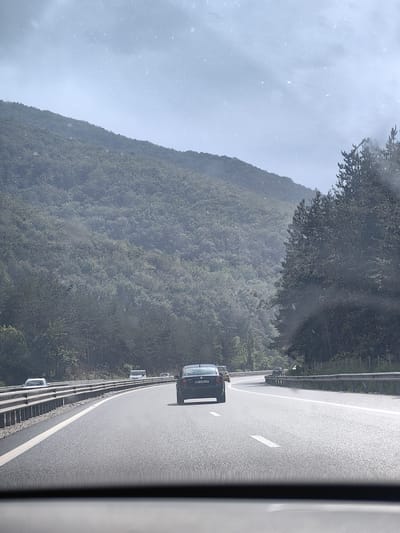 This media file depicts a view from inside a vehicle traveling on a highway in Churek, Bulgaria. The scene is characterized by a grey, overcast sky with visible particles, possibly snow or dust, suspended in the air. A lush, green, forested mountain range dominates the background. The highway is paved and marked with white lane lines, curving to the right. Several vehicles are visible on the road, including a dark-colored car in the foreground with a license plate reading "BP 3193 CP". Other cars and a white van are further down the highway. Metal guardrails line the side of the road. The overall impression is of a scenic drive through a mountainous, possibly rural, area under possibly inclement weather conditions. The time of day appears to be daytime, given the ambient light, but the heavy cloud cover creates a diffused and somewhat muted illumination.