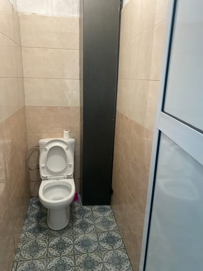 The image shows a toilet in a small bathroom. The toilet is white and made of ceramic. The toilet seat is up, and the toilet bowl is empty. The toilet is situated in a small bathroom with beige tile walls and a blue and white patterned tile floor. There is a tall, rectangular black panel to the right of the toilet, and another wall with a white door to the right of the panel. The bathroom is lit by overhead lighting, and there is no one in the room. The image was likely taken in a public restroom, but there are no other details available.