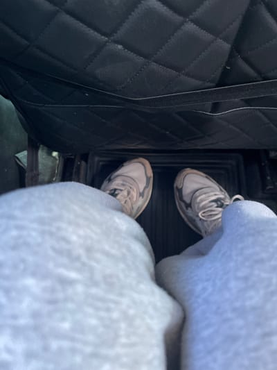 The image shows a person's legs and feet inside a vehicle. The person is wearing light gray sweatpants and white sneakers with dark gray accents. The feet are positioned on a black floor mat. Behind the feet, the interior of the vehicle is visible, including a quilted black seat or panel. The scene appears to be indoors, possibly in a parked vehicle. There is no discernible information about the city of Марьевка, Russia, within the image itself.