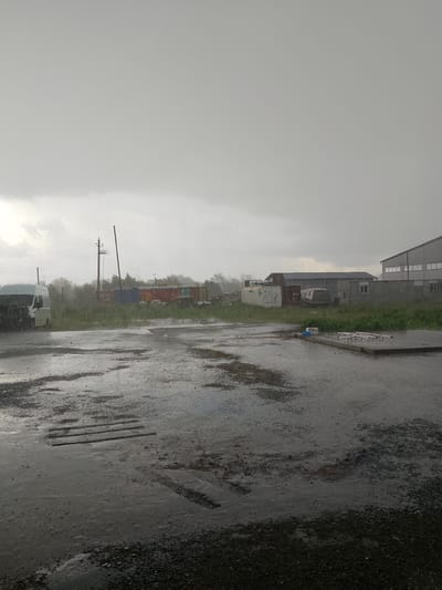 An outdoor, medium shot captures a rainy day scene in Мичурин ауылдық округі, Kazakhstan. The foreground is a wet, dark asphalt surface with puddles reflecting the overcast sky. Several rectangular, raised wooden planks are visible partially submerged in the water. In the mid-ground, a white van is parked on the left, with visible streaks of rain falling around it. Beyond the van, a grassy area is lined with colorful shipping containers, including red, blue, yellow, and white. Electrical poles and wires are present. In the background, industrial buildings with grey roofs and white walls are situated. The sky is a uniform, dull gray, indicating heavy cloud cover and ongoing rain. No people are visible in the scene.