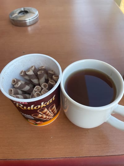 This media file depicts a close-up, overhead view of a wooden table with various items. The primary subjects are a white ceramic mug filled with dark liquid, likely tea or coffee, and a container of wafer roll snacks. The mug is positioned on the upper right side of the frame, and the container of snacks is below and to the left of the mug. The container appears to be made of cardboard and is adorned with branding that includes images of the wafer rolls and text. Some of the wafer rolls are visible spilling out of the container. To the far left of the frame, a small, circular metal object, possibly an ashtray or a small tin, is present. The lighting suggests it is daytime, with natural light illuminating the scene. The overall impression is of a casual break or snack time, likely indoors or in a sheltered outdoor area given the presence of the mug and snacks. There is no visible text that is legible for identification. The setting is a simple wooden surface, with hints of a darker, possibly red border visible on the right edge of the frame. The scene is static, with no discernible activity beyond the arrangement of the objects.