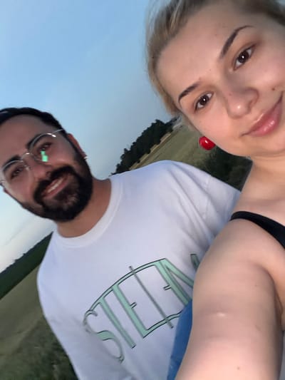 A selfie shows a man and a woman outdoors, with a blurry background of a field and trees. The man, on the left, wears glasses and a white t-shirt with a light green logo. He has a beard and is looking at the camera with a slight smile. The woman, on the right, has blonde hair and is also looking at the camera, smiling. She is wearing a black top and has large red earrings shaped like cherries. The sky in the background is a pale blue, suggesting it is either early morning or late evening. The scene appears to be in a rural or suburban area, possibly on the outskirts of Oberjettingen, Germany, given the open fields and tree line.