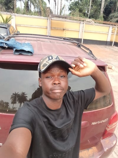 A young male subject, wearing a black t-shirt and a camouflage-patterned baseball cap displaying "DA" on the front, takes a selfie. His right hand is raised, grasping the brim of his cap. He stands in front of the rear of a dark red Toyota Highlander SUV, which has "HIGHLANDER" embossed on its tailgate. A pair of blue denim jeans are draped over the vehicle's roof rails. In the background, a light-colored perimeter wall with darker vertical accents is visible, beyond which tall palm trees and dense green vegetation extend upwards under an overcast sky. A partial view of another dark vehicle is present on the far left. The ground on the right side of the frame appears unpaved. The scene is located in Itu, Nigeria.