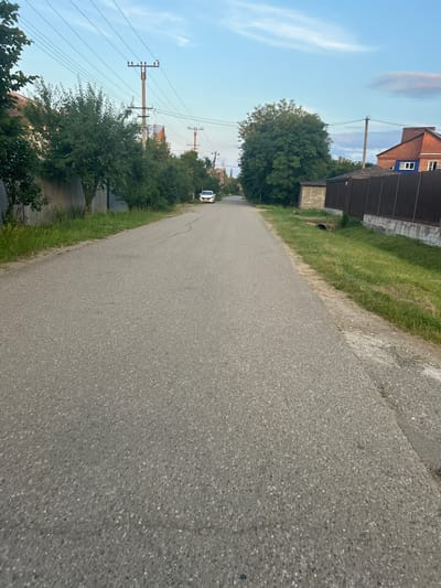 This is a wide-angle, low-angle shot of a paved road curving to the left, with a clear sky and some wispy clouds above. The image is oriented vertically. On the right side of the road, there is a grassy verge. Beyond the verge, on the left, are several elements of a rural or suburban setting.
In the foreground and midground, there is a dark brown, shed-like structure adjacent to a building with a reddish-brown brick facade. The building has a dark roof and several windows. A large, leafy green tree stands between the road and the buildings. Further down the road, more trees and vegetation line the left side, behind a grey metal fence. Power lines and utility poles are prominent in the background, stretching across the sky. A white car is visible on the road in the distance.
The lighting suggests it is daytime, likely late afternoon or early evening, given the soft light and the presence of clouds. The overall scene depicts a quiet, rural road in what appears to be a residential area. There are no people visible in the image. The mood is peaceful and serene. No text is visible in the image.