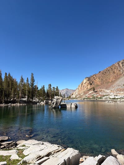 The image depicts a serene mountain lake scene in Mineral King, United States, under a clear, bright blue sky. The foreground is dominated by large, light-colored granite boulders with patches of green grass. The lake itself is a deep, clear turquoise, with visible rocks and pebbles beneath the surface in the shallower areas. In the mid-ground, several large, irregularly shaped rocks rise from the water, creating a focal point. The left side of the lake is bordered by a dense stand of tall, dark green pine trees. The right side of the lake is framed by a steep, rocky mountainside with reddish-brown hues, sparsely populated with small trees and shrubs. Further in the distance, additional mountains are visible, their peaks slightly softened by atmospheric perspective. No people or animals are present in the scene. The overall impression is one of natural beauty and tranquility.
