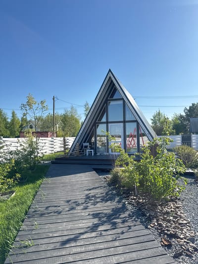 The image captures a scene in Pad', Russia. The focal point is a triangular-shaped building with a black and white frame, featuring a large glass facade. Through the glass, red scaffolding-like structures are visible inside. The building has a dark wooden deck. The ground adjacent to the deck is covered with gravel and vegetation. The sky is clear and blue. Power lines stretch across the upper left portion of the frame. In the background, there appears to be another structure, possibly a building or an elaborate fence. Also visible is a tall wooden pole with attached electrical equipment.