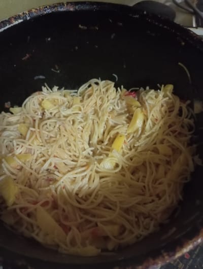 A close-up, overhead shot shows a black pot filled with cooked spaghetti noodles mixed with sliced yellow vegetables, possibly potatoes or bell peppers, and small pieces of red ingredients, which could be tomatoes or chili peppers. The noodles are thin and appear to be a light golden color, some coiled and others in strands. The yellow vegetables are cut into irregular, bite-sized pieces and are interspersed throughout the pasta. The red elements are small and scattered, adding pops of color. The pot itself is dark and shows signs of use, with some residue or staining around the rim. The overall lighting is dim, suggesting the image was taken indoors, possibly in a kitchen setting. The focus is entirely on the food within the pot. There is no visible text in the image. The context provided is Nurabad, Uzbekistan, implying this is a dish prepared or consumed in that location. The image captures a moment of food preparation or serving.