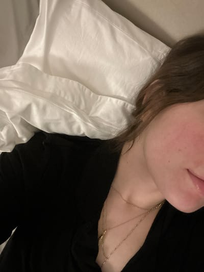 A person rests on a white pillow, a portion of their face visible. They wear a black top. A layered gold necklace adorns their neck. The pillowcase appears smooth and slightly creased. The person's skin is fair. Their lips are slightly parted. The scene is dimly lit, casting soft shadows.