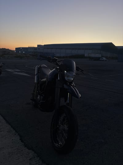 The image displays a static outdoor scene captured at dusk in Ypsonas Municipality, Cyprus. A dark-colored, supermoto-style motorcycle is centrally positioned in the foreground, facing towards the viewer's right. Its front headlight unit is distinctly visible, with internal reflective elements catching faint light. The motorcycle is largely silhouetted against the ambient light, obscuring finer details but revealing spoked wheels. The ground consists of a dark asphalt surface with visible tire marks, suggesting previous vehicle activity, and a white painted line along the left edge. In the midground, a white tanker truck is parked, partially obscured by the motorcycle on the left, with a small white passenger car parked behind it to the right. The background is dominated by a series of large, grey industrial-style buildings featuring multiple windows. The sky above presents a gradient from a warm golden-orange hue near the horizon to a pale blue towards the top of the frame, indicative of twilight. Sparse, dark vegetation and unkempt terrain are visible along the distant left horizon. No people are present, and no active movements or interactions are depicted.