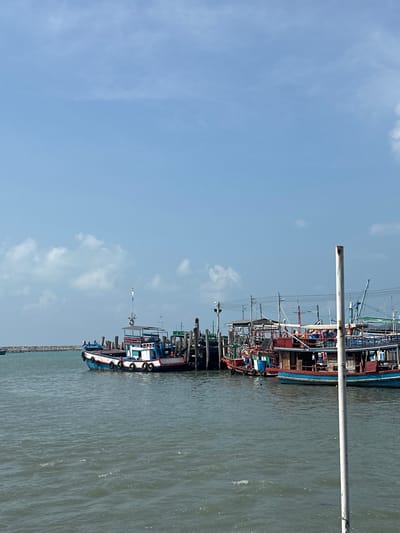 A vertically oriented photo shows boats moored at a pier. The sky is light blue with scattered clouds. The water is a dull grayish-green. Fishing vessels of varying sizes are tied along the pier. The pier extends into the water. A tall metal pole is also visible. The image captures the atmosphere of the coastal town of Ban Phe, Thailand.