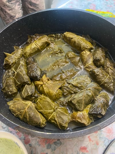 A top-down, close-up shot shows a pot of stuffed grape leaves, a dish known as dolma. The grape leaves are a deep green, some appearing wilted and darker, while others are a lighter, more vibrant green. They are tightly rolled and packed closely together in the pot, submerged in a light-colored liquid. The pot itself is black and appears to be made of a non-stick material, with a slightly textured interior. In the bottom left of the frame, a white bowl is partially visible, containing a pale green, creamy substance, likely a sauce or dip. The background is out of focus, but suggests a patterned tablecloth in shades of pink and white, and a person wearing camouflage-patterned pants. The image is well-lit, emphasizing the textures and colors of the food. This scene is not specific to Shiryaevo, Russia, and no elements within the image suggest this location.