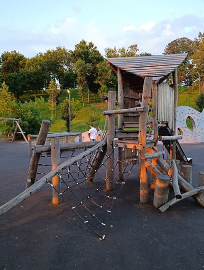 In a park setting within the city of Kelermesskaya, Russia, a wooden playground structure stands prominently. The playground features a net made of black rope stretched between wooden beams, a wooden tower with a sloped roof, and a slide. To the left of the main structure, a swing set is visible. In the background, a grassy hill is dotted with trees. Two individuals, a man and a woman, are standing near the playground, looking towards it. The ground around the playground is a dark, textured surface. The lighting suggests it is either early morning or late afternoon, with warm light casting shadows.