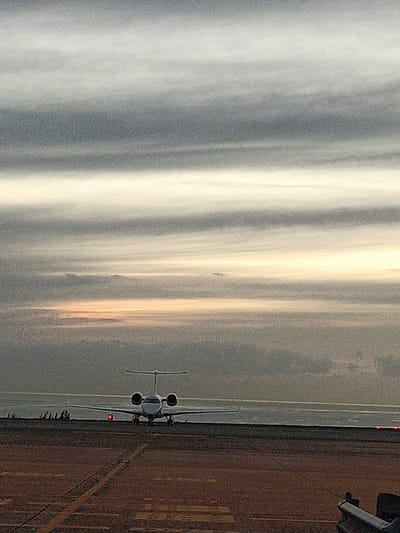 A private jet sits on a runway, facing the camera head-on. The background features a sky filled with horizontal streaks of gray and white clouds, suggesting either dawn or dusk. The horizon is faintly visible. Runway markings are evident on the foreground. Some ground equipment is visible in the lower right. The overall atmosphere is subdued and possibly overcast.