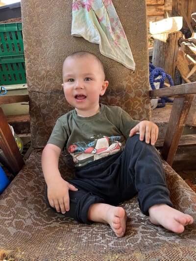 A young boy sits on a weathered armchair outdoors, in what appears to be a rural setting, possibly a yard or a rustic porch. The boy is wearing a green t-shirt with a graphic print and black pants. His feet are bare and resting on the seat of the chair. He has a curious expression with his mouth open. The armchair itself is made of wood and upholstered with a patterned fabric, showing signs of age and wear. In the background, there are elements suggesting a somewhat unkempt outdoor space, including stacks of wood, a blue rope, and a green plastic crate containing what looks like soil or mulch. A piece of patterned fabric hangs from the back of the chair. The overall atmosphere is casual and informal. The location is identified as Аряж, Russia.