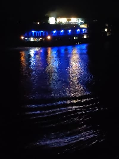 The image depicts a night scene in Tikhonovo, Russia, likely on a body of water. A multi-story structure, possibly a boat or building on the water's edge, is illuminated with blue and white lights. The lights reflect on the water's surface, creating a vibrant blue and orange shimmering effect. The foreground is darker, showing the texture of the water with slight waves. The overall atmosphere is dark and serene, punctuated by the bright, colorful reflections.