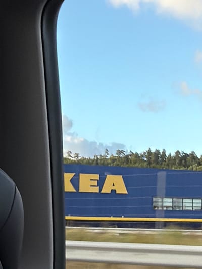 The image shows a bright blue IKEA building with large yellow letters spelling "KEA" on its side. The building has a yellow stripe running along its base. Above the building, a line of green trees is visible against a pale blue sky with scattered clouds. The foreground is occupied by a dark gray car interior, suggesting the photo was taken from inside a vehicle. The scene is outdoors, and the lighting indicates it is daytime. Based on the provided information, this location is in Ödsmål, Sweden.