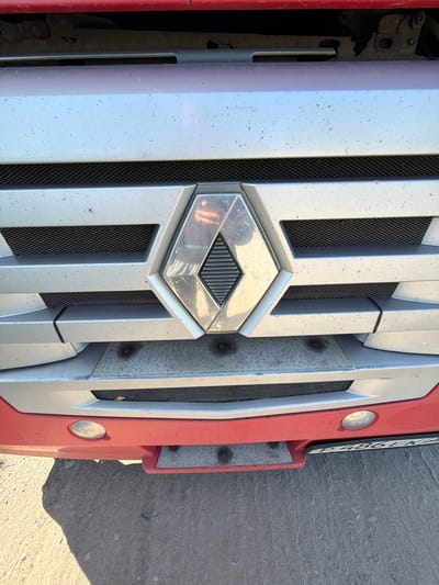 The image shows the front grille of a red and silver Renault truck, parked on a dirt and concrete surface. The truck's logo, a diamond shape with a horizontal line through it, is prominently displayed in the center of the grille. There are two circular headlights visible on the lower part of the bumper. A license plate with alphanumeric characters is affixed to the front of the truck. The environment appears to be outdoors, possibly in a rural or industrial area, as suggested by the ground surface and the presence of a commercial vehicle. The lighting indicates it is daytime, with a shadow cast to the left of the truck. The specific location in Zarech'e, Russia, cannot be determined from the visual information alone.
