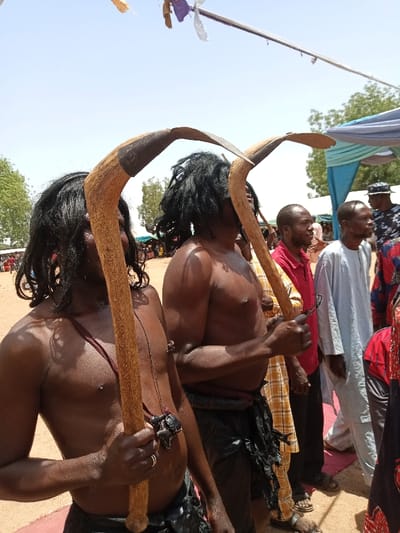 The scene takes place outdoors in a community gathering in Zum, Nigeria (latitude: 10.116, longitude: 13.046). Two shirtless men are standing in a line of other men, holding large, curved wooden staffs with metal tips. The man on the left has his staff resting against his shoulder and is wearing a thin, dark strap around his neck with a small, beaded pendant hanging from it. He is wearing a dark belt with a large, black object attached to it. The man on the right is holding his staff in front of him with his right hand. He is also wearing a dark belt. Both men are wearing black fabric wrapped around their waist and legs. They have long, black hair, and their skin is tanned from the sun. The men behind them are wearing various colored clothing, including plaid shirts, long-sleeved white shirts, and a blue and white patterned robe. The ground is dry and dusty. The setting suggests a social or celebratory occasion.