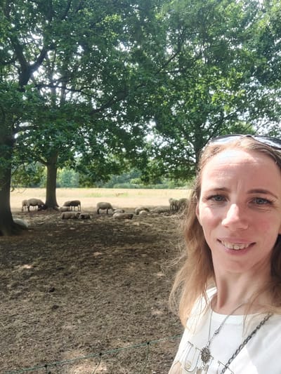 A woman with light brown hair and blue eyes, wearing sunglasses perched on her head and a white t-shirt with two necklaces, smiles at the camera. She is standing in a grassy area with scattered trees, likely in the vicinity of Isselburg, Germany, given the contextual information. In the background, a flock of sheep grazes and rests under the shade of the trees. The ground is covered in dry grass and dirt.