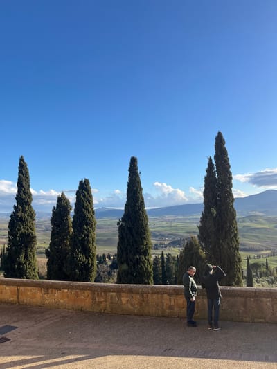 The image, oriented correctly, displays a picturesque panoramic view of the Tuscan countryside under a clear blue sky. In the foreground, on the right side of the frame, a stone retaining wall or balcony provides a vantage point. Two men are standing on the paved surface behind this wall. The man closer to the viewer wears a green puffer jacket and dark pants, gazing out at the landscape. Further back, a second man in a dark coat and dark pants holds a phone or camera up, appearing to photograph or video the scenic vista.

A distinctive line of tall, slender cypress trees, characteristic of the region, runs diagonally from the bottom-left to the mid-right, separating the immediate foreground from the sprawling landscape beyond. These trees stand out against the vibrant blue sky, which features scattered white clouds. The background is dominated by gently rolling hills and valleys, showing a patchwork of green fields and cultivated land, stretching towards distant mountains or larger hills under a bright sun. The scene suggests a pleasant, sunny day, likely late morning or early afternoon, offering an expansive view from an elevated position in Pienza, Italy. There is no visible text in the image.