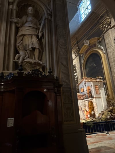 The image depicts the interior of the Ferrara Cathedral, Ferrara, Italy. The scene is characterized by dim illumination, with natural light entering from a high, arched window visible in the upper right quadrant, casting a blue hue.
In the left foreground, a dark stained wooden confessional booth is present, featuring carved ornamentation along its upper edge, a recessed archway, and a small white circular object affixed above the arch. A printed paper displaying a cross symbol is attached to the left side of the confessional.
Vertically aligned above the confessional, a light-toned marble statue occupies an ornate recessed niche within a structural pilaster. This statue portrays a robed male figure with an outstretched right arm, palm open, and his left hand grasping an unidentifiable object. A smaller, nude putto figure is positioned at the base of the male figure, gazing upwards. An inscribed pedestal supports the main statue, though the text is illegible.
To the right, a large, illuminated Nativity scene (presepio) is visible. It comprises a multi-story architectural model with a straw-covered roof and windows emitting a warm yellow light. Within the scene, multiple figures, including human and animal forms, are arranged. A low blue-colored barrier delineates the perimeter of the Nativity scene from the patterned floor. The background features additional ornate architectural details, including decorative pilasters, arched alcoves, and what appears to be a dark painting within one such alcove.