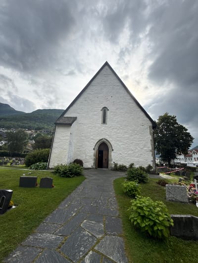 The image shows the exterior of the Aurland Church in Aurlandsvangen, Norway, on an overcast day. The church is a white, stone building with a steep, dark-tiled roof and a large, triangular facade. An arched wooden doorway with visible details of the interior is centrally located on the facade, and a small, arched window is positioned above it. A paved pathway made of irregular dark stones leads from the foreground to the church entrance.
The church is surrounded by a graveyard with several headstones visible on the grassy grounds. To the left of the church, a lush green hillside with houses and trees slopes upwards, indicating the town's setting. A large, mature tree stands to the right of the church, and in the background, more houses and the distinctive Norwegian landscape of mountains and fjords are partially obscured by the clouds and the tree. Red and white caution tape is visible in the distance, potentially indicating a restricted area or ongoing work. No people are visible in the image. The overall atmosphere is somber due to the cloudy sky.