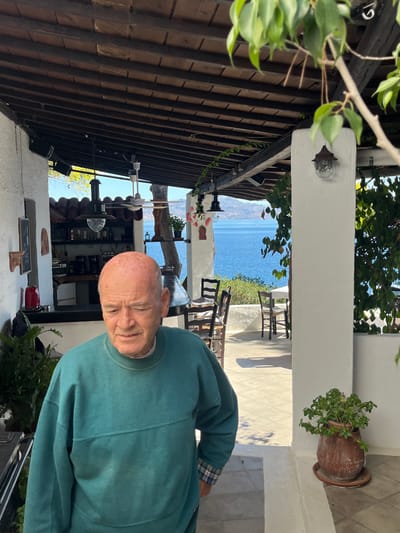 The image depicts an elderly bald man with fair skin and red blotches on his scalp, wearing a teal-green long-sleeved sweatshirt over a plaid shirt, standing on a tiled patio in what appears to be an outdoor restaurant or cafe. His gaze is directed downwards and slightly to the left. The scene is set in Megalochori, Greece.

To the man's left and behind him, a white counter area with bottles and a red kettle suggests a bar or service station. Above, a wooden beamed ceiling with white supporting structures extends over the patio. Several light fixtures and a ceiling fan are visible.

Beyond the patio, the deep blue Aegean Sea is prominently featured under a clear sky, with distant landmasses and a faint sailboat visible on the horizon. The patio itself is furnished with dark-colored chairs and tables, some of which are partially obscured by white pillars. Green foliage and potted plants are strategically placed throughout the area, including a large potted plant on the lower right foreground. Strong sunlight indicates a bright day.