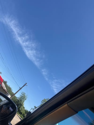 The image shows a view of the sky from inside a vehicle. The sky is blue with a long, thin cloud formation stretching across the upper part of the frame. Utility poles and power lines are visible in the lower right quadrant, alongside trees. A vehicle's side mirror is visible in the foreground, reflecting a hand holding a smartphone. In the background, a red vehicle and building structure can be seen partially. Based on the context and provided location, the scene is likely in the area of Topchii, Bulgaria.