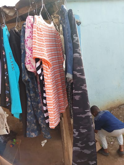 The image displays an outdoor scene, likely in Ohokobe Ndume, Nigeria, centered around a collection of clothing hanging on a makeshift rack. Numerous garments are suspended from simple wire hangers, suggesting either clothes drying or items for sale. Visible clothing includes a bright turquoise long-sleeved top with the text "OH, YES!", a black shirt, a sheer pink and white patterned fabric, a dark blue floral patterned garment, an orange and white striped top, a black and white striped top, blue denim jeans, and a black and grey camouflage patterned fabric. A white lace bra is partially visible on the far left. The rack itself appears to be constructed from rough wooden planks, with a corrugated roof visible above.

In the lower right corner, a person, possibly a child or young man, is partially obscured by the hanging clothes. He is seen from behind, crouching down, wearing a blue short-sleeved shirt, light-colored pants, and dark slides or sandals. The background features a light blue-painted wall. The ground is unpaved dirt. To the left, a small wooden stool or box holds several colorful plastic bottle caps (green and orange). A crumpled white item and a cardboard box are also on the ground near the crouching figure. The natural light indicates it is daytime.