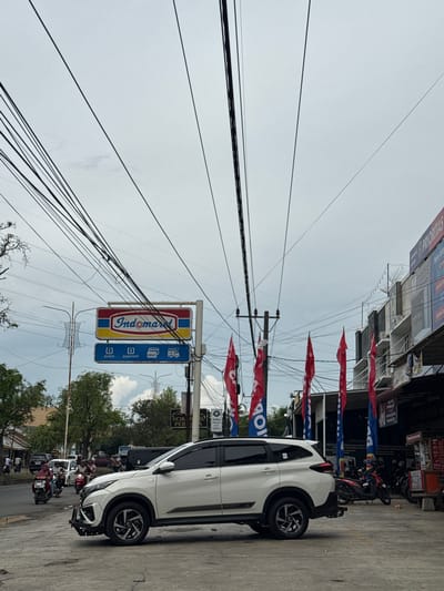 In the city of Meunasahmanyet, Indonesia, a white SUV is parked on a street with a cloudy sky overhead. The vehicle, positioned facing right, is partially obscuring a sidewalk with a row of shops. The shops feature red and blue flags, and signs indicate services like "pulsa" (phone credit), "payment", "DEBIT", and "PAKET" (package delivery).

Behind the SUV, a prominent "Indomaret" convenience store sign is visible, mounted on a pole. The street is active with traffic, including motorcycles and other vehicles. Trees and utility poles with numerous overhead wires are scattered throughout the scene, contributing to the urban landscape. The ground is paved with concrete and asphalt. The weather appears overcast.
