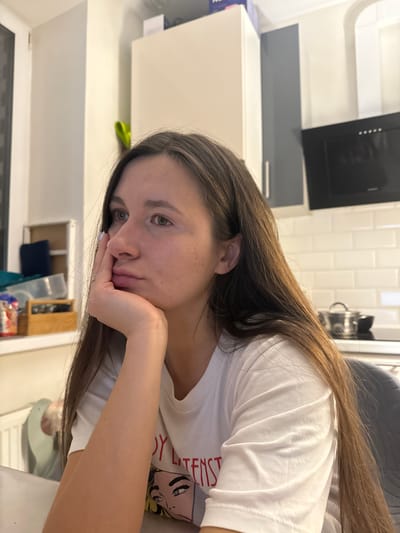 A female individual is depicted indoors, seated at a table within a residential kitchen or dining area in Осоргино, Russia. The subject, possessing long brown hair and fair skin, rests her left hand, adorned with a three-band ring on the ring finger and white nail polish, under her chin. She wears a white short-sleeved t-shirt featuring a partial graphic design. Her gaze is directed towards the left side of the frame. The background reveals a kitchen area with white subway tile backsplash, a black range hood, white and light grey cabinetry, and a metal pot with a lid on a stovetop. To the left, a window or door frame, a white radiator, and a shelving unit containing various items including a blue container and a wooden box are visible.