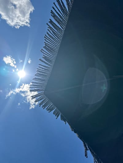 An extreme low-angle outdoor shot captures a dark, fringed textile against a bright blue sky. The sun is prominently visible in the upper left quadrant, creating a lens flare that extends across the frame, with a faint green cross shape visible within the flare's circular reflection. A few white clouds are scattered across the sky. The textile, which appears to be draped or hung, has a dense fringe of numerous small, dark, looped strands along its visible edge. The overall impression is of looking up from beneath the textile towards the sky on a sunny day, possibly in the urban environment of Γεωπονικά, Greece, where such textiles might be found outdoors.