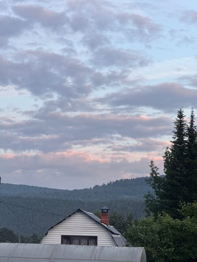 The image shows a rural scene with a house, a forest-covered mountain range, and a cloudy sky. The house, located on the right side of the frame, has a white, clapboard exterior, a dark grey roof, and a red brick chimney. A window with dark panes is visible on the side of the house. In the foreground, to the right, a portion of a grey, translucent structure, possibly a greenhouse or shed, is visible. Behind the house, a dense, dark green forest covers a mountain range that stretches across the middle ground. The sky is filled with wispy, grey clouds, with hints of pink and orange suggesting either sunrise or sunset. The overall mood is tranquil and serene. The time of day appears to be either early morning or late evening. The location context is Ovsyanka, Russia. There is no visible text in the image.