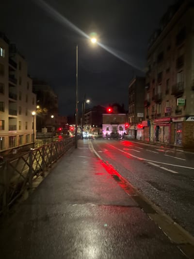An evening scene of a city street. Rain reflects red light from traffic signals and buildings. The wet pavement glistens, delineating lane markings. Buildings line the left, including several structures with multiple stories and lit windows. A streetlight shines brightly in the dim sky. A metal structure extends along the right edge of the scene. The photo is tilted slightly, giving a distorted perspective of the urban environment.
