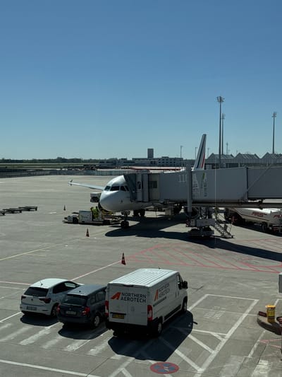 An overhead view captures an airport apron scene under a clear, bright blue sky with strong sunlight casting distinct shadows. An airplane, possibly an Airbus A320, is parked at a gate, connected to a jet bridge.
The jet bridge displays the gate number "116". Ground support operations are evident around the aircraft. A white refueling truck with "AFSA" visible on its side is positioned behind the jet bridge. Near the front of the aircraft, other ground equipment, possibly a tug, is present.
On the tarmac, several vehicles are parked. A white van with "NORTHERN AEROTECH" branding is prominent, displaying the license plate "DS 78 588" and a Peugeot logo. Next to it, a dark-colored car with license plate "FS IRU 121" and a white car with license plate "OF DE 120" are visible. Orange traffic cones are placed on the ground, and a yellow "F.O.D." container is also present.
The asphalt features clear markings, including red and white lines, and a designated area with the text "AIRPORT DELIVERY." A no-parking sign is also visible. The backdrop includes parts of the airport terminal buildings and distant trees along the horizon. The scene depicts typical daytime ground activity at an airport in Oberding, Germany.