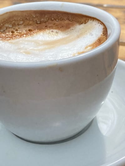 A close-up shows a white ceramic cup partially filled with a beverage, likely coffee, with a foamy top layer. The cup rests on a white saucer. The scene is photographed with shallow depth of field, suggesting an indoor setting. There are no people visible. The location and date are not identifiable from the image. There is no mention of Krapina or Montenegro.
