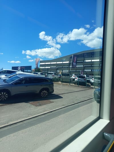 The image depicts a car dealership lot in Laxou, France, on a sunny day with a bright blue sky and scattered white clouds. In the foreground, a dark grey SUV is parked, facing left. Behind it, several other cars are lined up, including white and silver models. A modern, multi-story building with large windows and a metallic facade serves as the backdrop. Banners with the "l'okasion" logo are visible on flags and the building. The scene is viewed from inside a vehicle, with a window frame partially obscuring the view on the right side. The ground is paved, with a metal grate visible in the foreground.