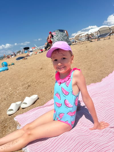 The media file shows a young girl lying on a pink striped towel on a sandy beach. She is wearing a blue swimsuit with a watermelon print and a pink ruffled collar. She is also wearing a purple hat. Her left arm is extended, touching the towel. Next to her are two white slides. The background shows a blurred view of other beachgoers, umbrellas, and the sea under a clear blue sky. The sun appears to be shining brightly, suggesting it is daytime. The text "be melon" and "wabe melon" (likely a misspelling of watermelon) is visible on the swimsuit. The location context indicates this is Blagoveschenskaya, Russia. The girl appears happy and relaxed.