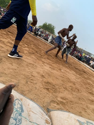Two men are wrestling in a sandy arena in Sokoto, Nigeria (13.049, 5.238). The man on the right is wearing blue shorts and is attempting to throw the man on the left, who is wearing a brown loincloth. The man on the right has his left arm wrapped in white tape. The man on the left is holding his left arm in front of his chest. There are spectators seated around the edge of the arena. The ground is sandy and there are white sacks bordering the arena. The photograph is taken from a low angle, looking up at the wrestlers. A person's foot is partially visible in the foreground of the photo. The man taking the photo is wearing blue pants and a black and white Adidas shoe. The man's pants have a green logo with the words "Sokoto" on it.